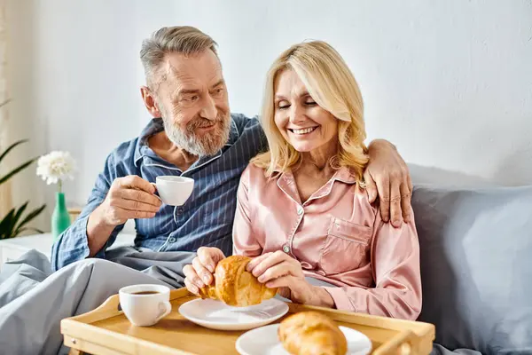 Una pareja amorosa madura en ropa de casa acogedora sentada en un sofá, saboreando el café juntos en un entorno cálido e íntimo. - foto de stock