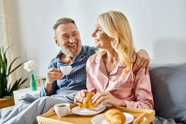 Una pareja madura y cariñosa está sentada en un sofá, disfrutando de una bandeja de comida juntos en su acogedora ropa de casa en el dormitorio. - foto de stock