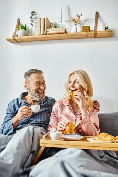 Un hombre y una mujer maduros disfrutan de una comida mientras están sentados en un cómodo sofá en su acogedor dormitorio, vestidos con ropa de casa. - foto de stock