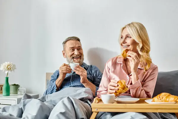 Pareja madura en ropa de casa sentada en un sofá, comiendo felizmente comida en un ambiente acogedor. - foto de stock