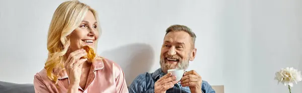 A mature loving couple sitting at a table, enjoying a meal together in their cozy homewear in the bedroom. — Stock Photo