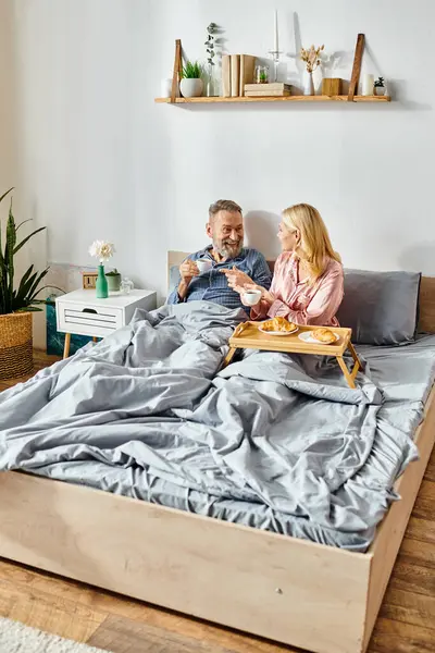 A mature loving couple in cozy homewear sitting closely together on a bed in their bedroom, sharing a quiet moment. — Stock Photo