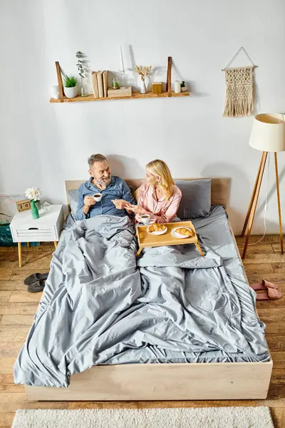 Una pareja madura y cariñosa en ropa de casa acogedora, sentados juntos en una cama, compartiendo un momento pacífico y tierno. - foto de stock