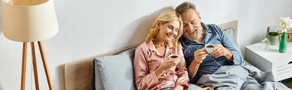 Una pareja amorosa madura en ropa de casa acogedora, sentada en una cama con café - foto de stock