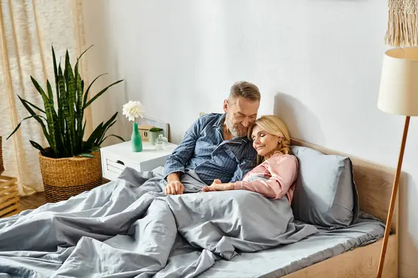 Un hombre y una mujer, en ropa de casa acogedora, abrazan amorosamente mientras que ponen en una cama debajo de una manta suave en su dormitorio. - foto de stock