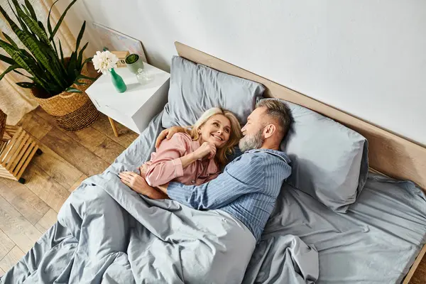 Una pareja madura y cariñosa en ropa de casa abrazándose y relajándose juntos en una cama, compartiendo un momento tranquilo de intimidad. - foto de stock