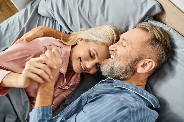 A mature loving couple in cozy homewear lay peacefully on a bed, sharing a tender embrace in their bedroom. — Stock Photo