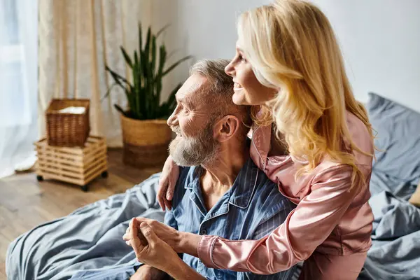 A mature loving couple in cozy homewear sit closely together on a bed, enjoying each others company in a quiet moment. — Stock Photo