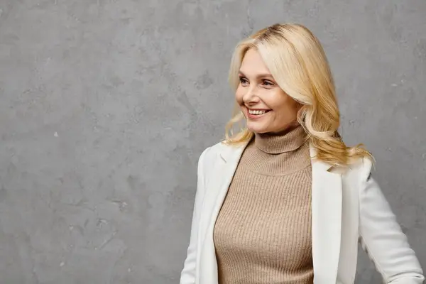 A mature woman exudes elegance in a tan turtleneck sweater and white blazer, striking a pose against a gray backdrop. — Stock Photo