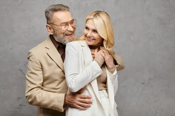 Un uomo e una donna maturi, elegantemente vestiti, in piedi vicino in un abbraccio amorevole su uno sfondo grigio. — Foto stock