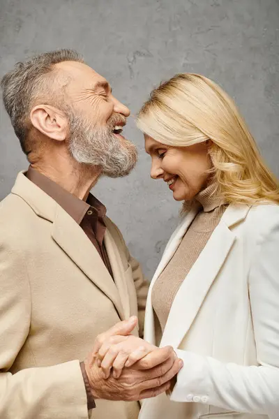 Elegant, mature, loving couple in debonair attires stand together against a gray backdrop. — Stock Photo