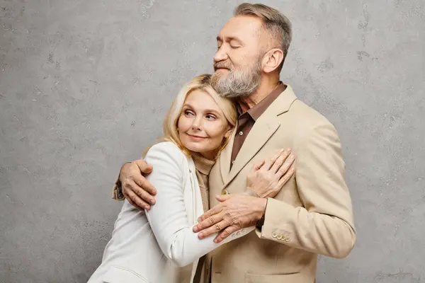 A mature loving couple in debonair attire warmly embrace each other in a graceful pose against a gray backdrop. — Stock Photo