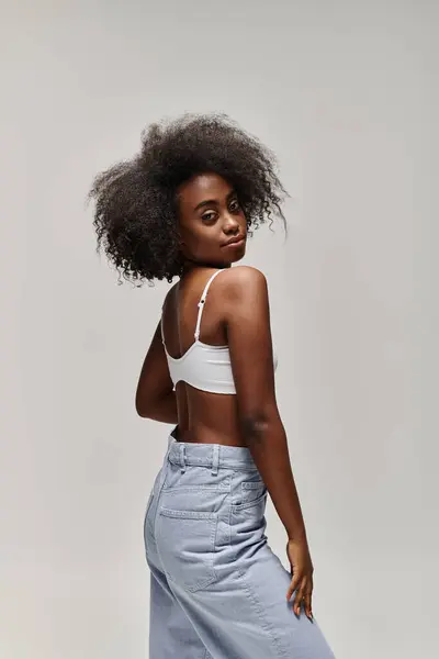 A beautiful young African American woman with curly hair dressed in a white top and blue pants exuding elegance and grace. — Stock Photo