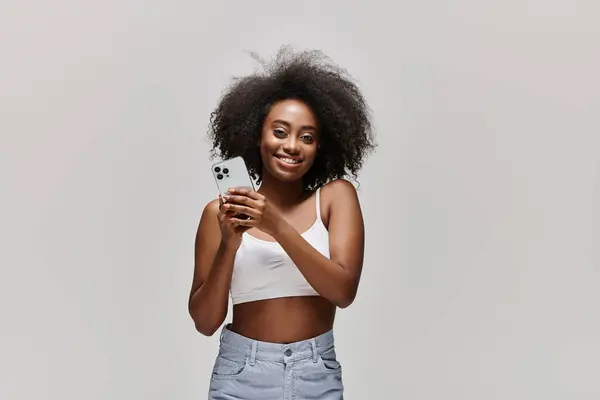 Une jeune afro-américaine aux cheveux bouclés tenant un téléphone portable dans ses mains, absorbée dans son écran. — Photo de stock