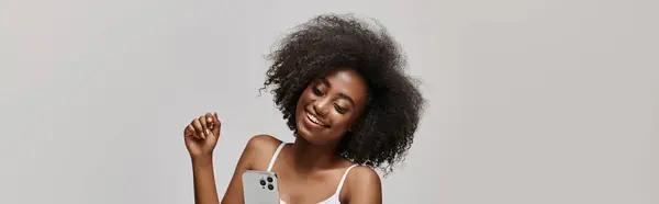 A beautiful young African American woman with curly hair holding a cell phone while wearing a white tank top. - foto de stock