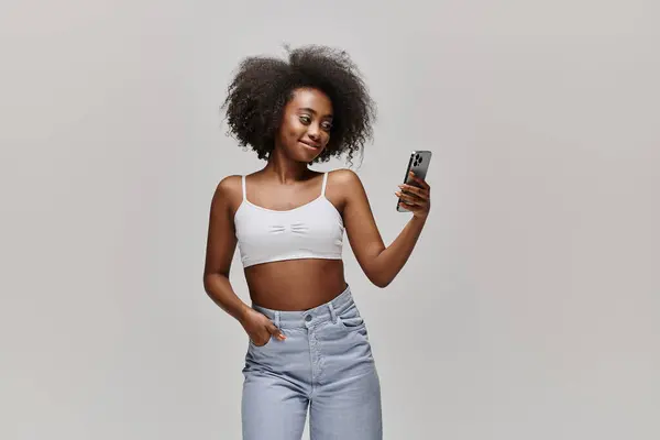Une femme afro-américaine élégante avec des cheveux bouclés portant un haut de culture, tenant un téléphone portable dans une pose à la mode. — Photo de stock