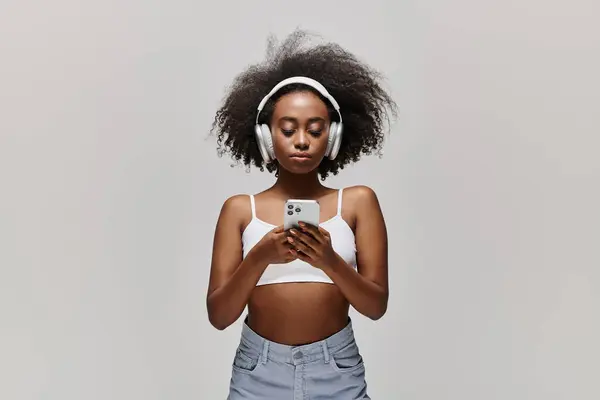 Une jeune afro-américaine aux cheveux bouclés, portant un casque, regarde attentivement son écran de téléphone portable. — Photo de stock