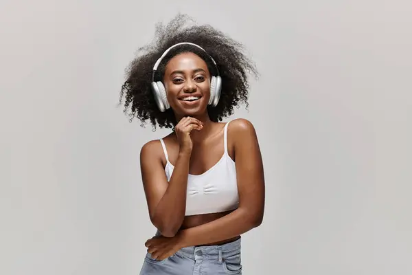 A young African American woman with curly hair stands in front of a white background, wearing headphones. — Stock Photo