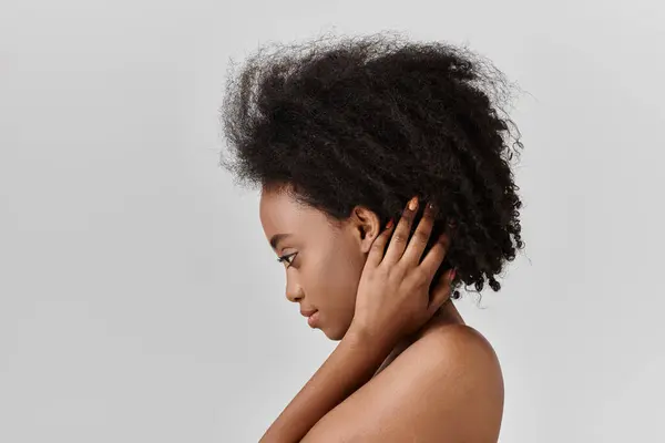 A beautiful young African American woman with curly hair gently rests her hand on her shoulder in a studio setting. — Stock Photo