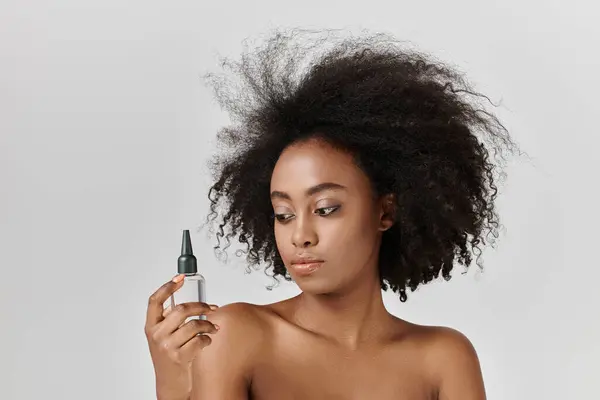 A young African American woman with curly hair holds a hair bottle, enhancing her beauty and emphasizing body care. — Stock Photo