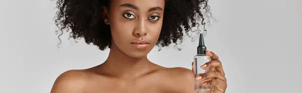 Une belle jeune femme afro-américaine aux cheveux bouclés tenant une bouteille de liquide dans sa main, concept de soins de la peau. — Photo de stock