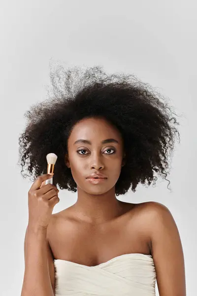 Joven mujer afroamericana con cabello rizado sosteniendo cepillo cosmético en un entorno de estudio que representa el concepto de belleza. - foto de stock