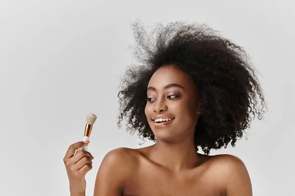 Una joven afroamericana con el pelo rizado sostiene un cepillo, centrándose en el cuidado de la piel y la belleza en un entorno de estudio. - foto de stock