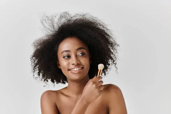 Eine schöne junge Afroamerikanerin mit lockigem Afro hält einen Pinsel in der Hand und strahlt Kreativität und Kunstfertigkeit in einem Atelierumfeld aus. — Stockfoto