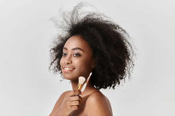 A confident young African American woman with curly hair holding makeup brushes in her hand. — Stock Photo