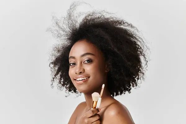 Афроамериканка с вьющимися волосами, держа щетки для макияжа, накладывает макияж в студии. — стоковое фото
