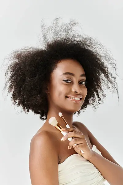 Красивая молодая афроамериканка с вьющимися волосами, держащая щетки для макияжа, воплощая концепцию ухода за кожей в студии. — стоковое фото
