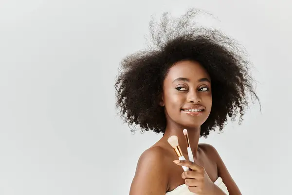 A young African American woman with curly hair holding makeup brushes in her hand in a studio setting. — Stock Photo