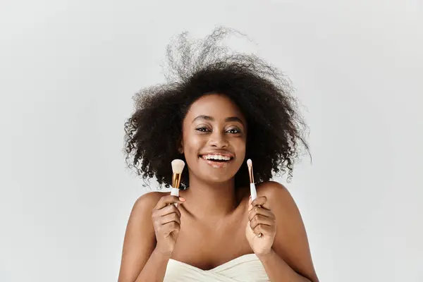 Una hermosa mujer afroamericana con el pelo rizado sostiene dos pinceles en sus manos mientras está de pie en un estudio. - foto de stock