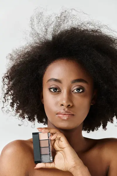 A young African American woman with a curly afro hairstyle holds a makeup palette, showcasing her skincare routine. — Stock Photo