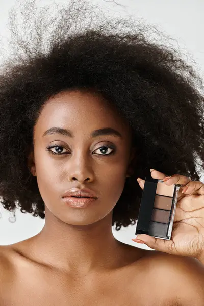 A young African American woman with curly hair holding a palette of makeup in a studio setting, emphasizing skin care. — Stock Photo