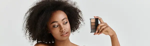 Une belle jeune afro-américaine aux cheveux bouclés dans un décor studio, tenant une palette de maquillage — Photo de stock