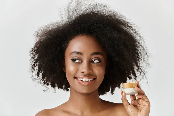 Una hermosa joven afroamericana con el pelo rizado sostiene un frasco de crema, encarnando la esencia del cuidado de la piel. - foto de stock