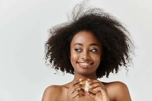 Uma jovem, bela mulher afro-americana com cabelo encaracolado segura frasco de creme, incorporando o conceito de nutrição e auto-cuidado. — Fotografia de Stock
