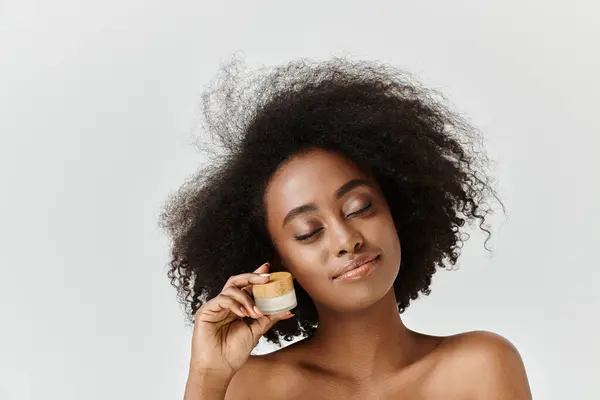 Una hermosa joven afroamericana con el pelo rizado sosteniendo un frasco de crema en su mano derecha. - foto de stock