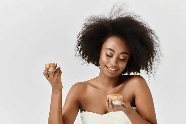 Una joven afroamericana impresionante, envuelta en una toalla, sostiene un frasco con crema - foto de stock