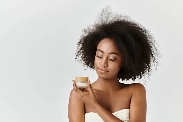 A young African American woman with curly hair stands in a towel, holding cream in studio setting. — Stock Photo