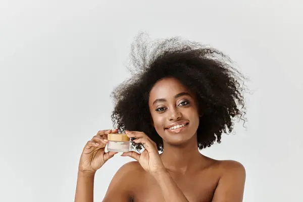 Una hermosa joven afroamericana sosteniendo una crema en un ambiente de estudio, haciendo hincapié en el concepto de cuidado de la piel. - foto de stock
