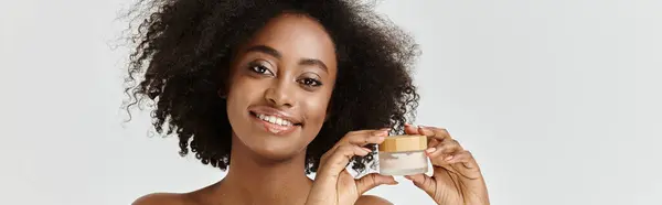 Красивая молодая афроамериканка с вьющимися волосами, держащая банку сливок в руке, продвигая уход за кожей. — стоковое фото