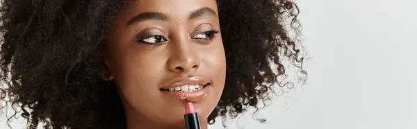 A beautiful, young African American woman with curly hair elegantly holds a lipstick tube in a studio setting — Stock Photo
