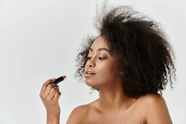 A beautiful young African American woman with curly hair applying lipstick, in a studio setting — Stock Photo