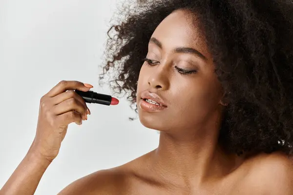A young African American woman with curly hair is skillfully applying lipstick to her face in a studio setting. — Stock Photo