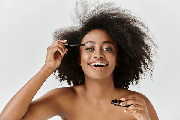 A beautiful young African American woman with her curly hair in a studio setting, applying mascara — Stock Photo