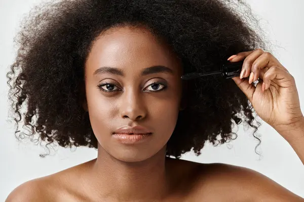 A beautiful young African American woman with curly hair gracefully applying mascara in a serene moment. — Stock Photo