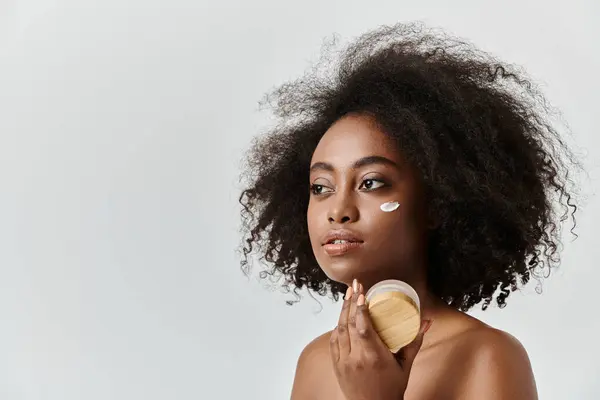 Eine junge Afroamerikanerin mit lockigem Haar hält sich ein Cremeglas vor das Gesicht und präsentiert ein Hautpflegekonzept. — Stockfoto