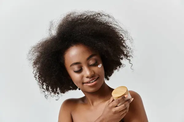 A stylish African American woman with a voluminous afro hairdo gracefully holds a cream in a studio setting. — Stock Photo